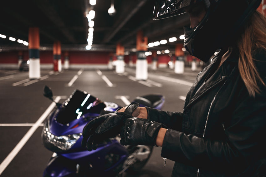 Plano lateral de una motociclista rubia con chaqueta negra y casco de seguridad poniéndose los guantes de cuero, preparándose para montar en una moto, de pie en un aparcamiento subterráneo. 