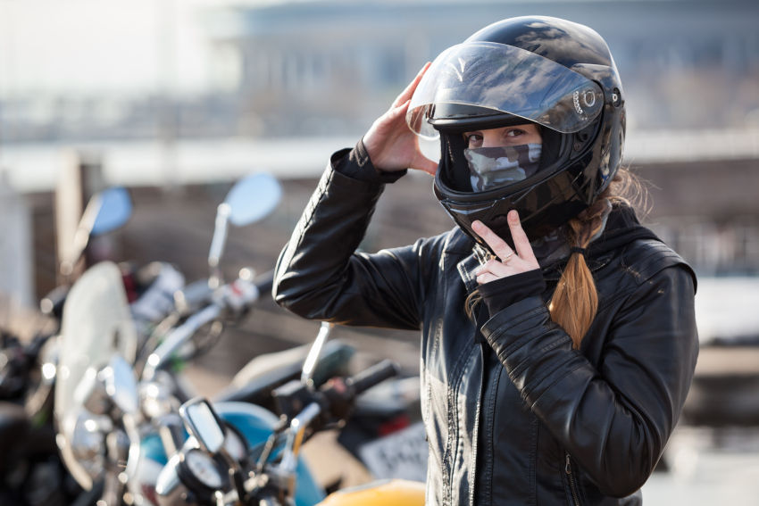 Chica de pie delante de su moto, sosteniendo su casco de protección. Va equipada además con chaqueta de cuero negra y mascarilla