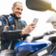 hombre con equipamiento motero subido en su moto aparcada, manejando su teléfono móvil