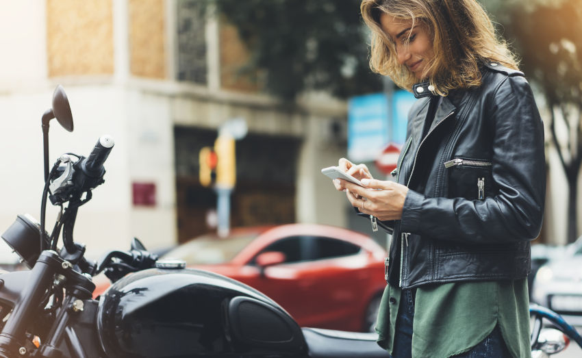 mujer mirando el móvil al lado de su moto aparcada en la acera de una ciudad