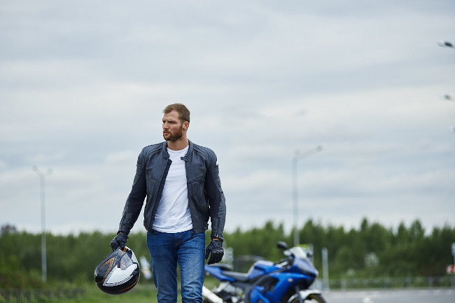 Hombre joven con casco en la mano y a su espalda la moto aparcada