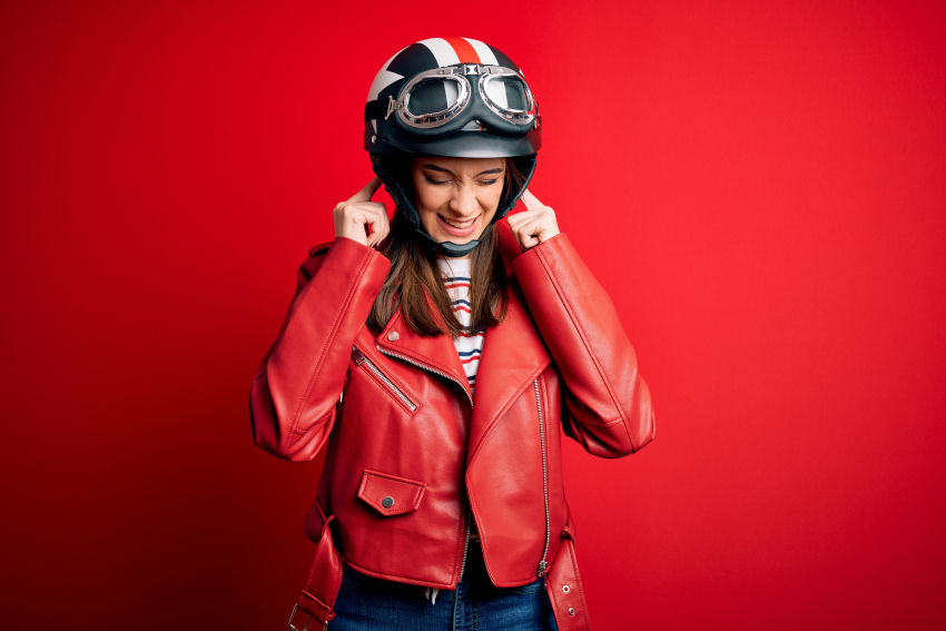 Chica con ropa de motorista (casco de moto y chaqueta roja) cubriendo sus oídos con los dedos, como expresión de molestia por el ruido.