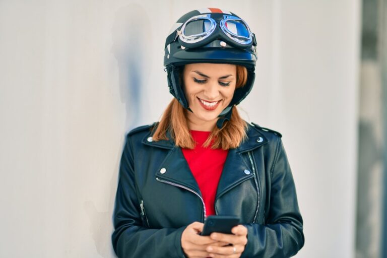 Mujer rubia equipada con chaqueta de cuero y casco y gafas de moto mirando al teléfono móvil que está sujetando entre sus manos