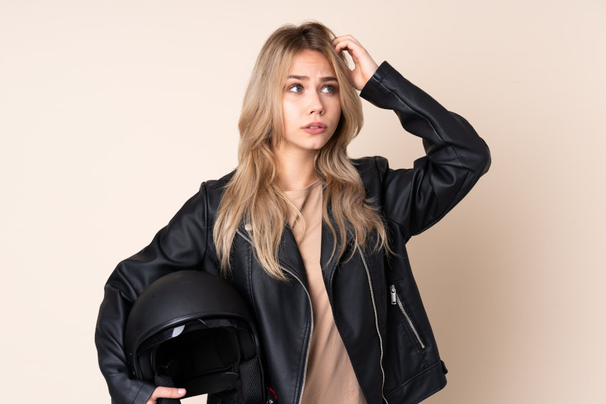 mujer joven vestida con chaqueta de cuero negra, rascándose la cabeza con una mano y sujetando un casco de moto en la otra