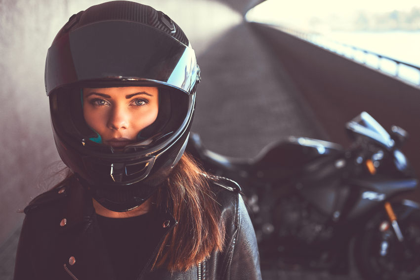 primer plano de una mujer con un casco integral de moto negro. En segundo plano se ve su moto de gran cilindrada aparcada.