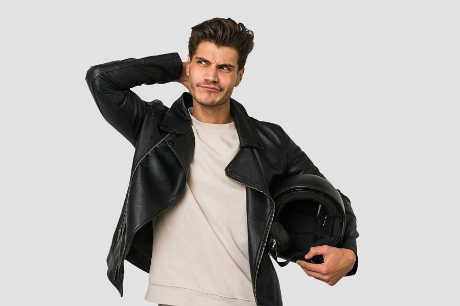 hombre vestido con cahqueta de cuero negra, sostiene un casco de motorista con una mano, mientras con la otra se toca la cabeza con expresión pensativa